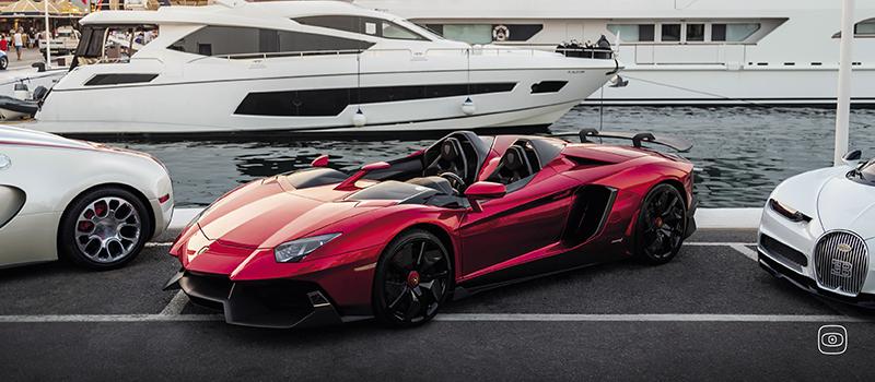 A red supercar with an open-top design is parked at a marina.